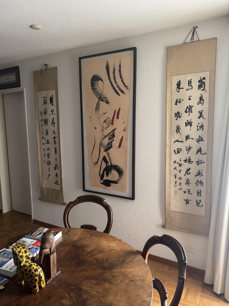Drei vertikale Wandrollen mit chinesischen Schriftzeichen und Kalligrafie hängen an einer weißen Wand über einem Holztisch.