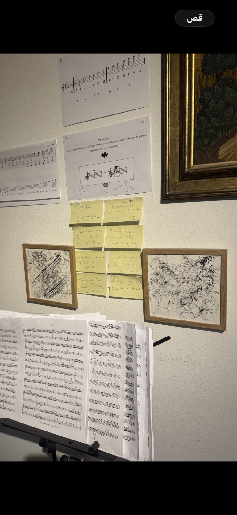 An einer weißen Wand hängen Notenblätter, ein erklärendes Blatt mit Musiktheorie, gelbe Haftnotizen und zwei gerahmte Skizzen.