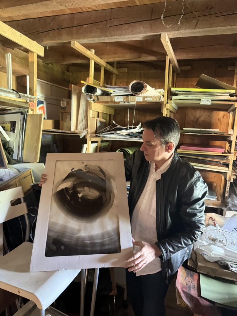 Ein Mann hält ein großes, abstraktes Schwarz-Weiß-Kunstwerk in einem mit Papier und Materialien gefüllten Holzwerkstattraum.