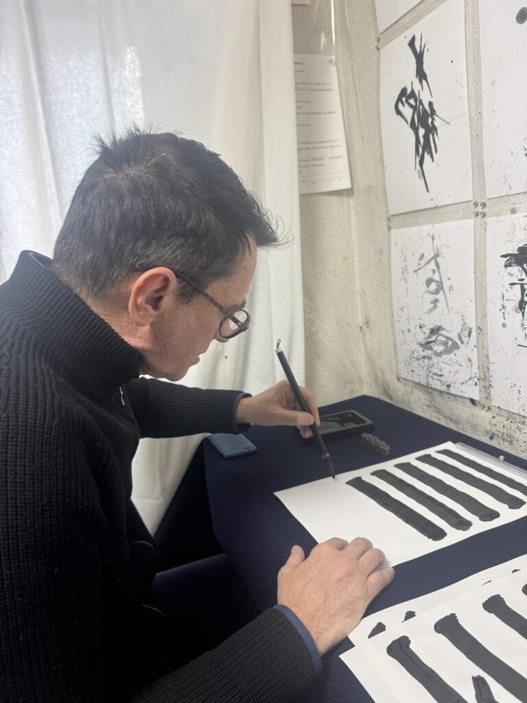 Ein Mann mit Brille schreibt mit einem Pinsel schwarze Linien auf ein weißes Blatt Papier an einem Tisch.