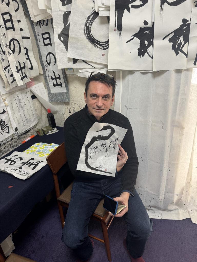 Ein Mann sitzt in einem Atelier und hält ein Blatt Papier mit schwarzer Kalligrafie in der Hand.