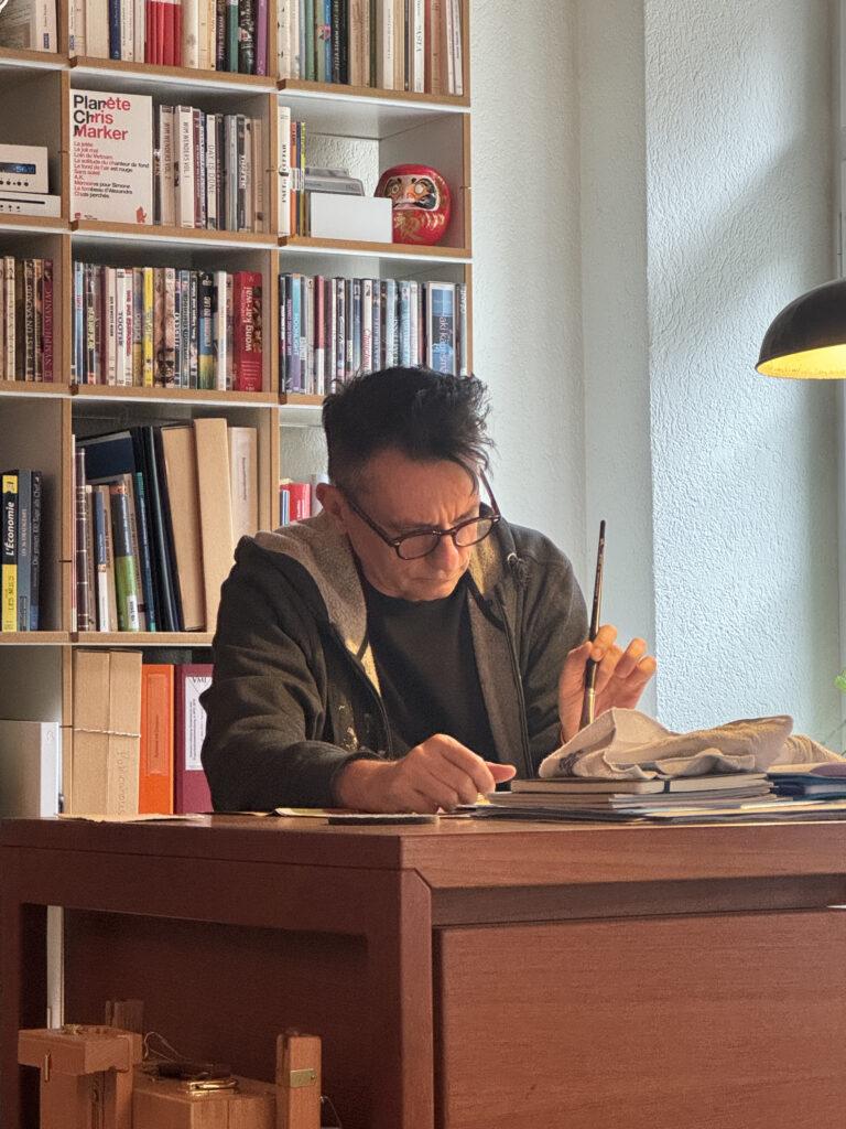 Ein Mann mit Brille sitzt an einem Schreibtisch vor einem gut gefüllten Bücherregal und schreibt.