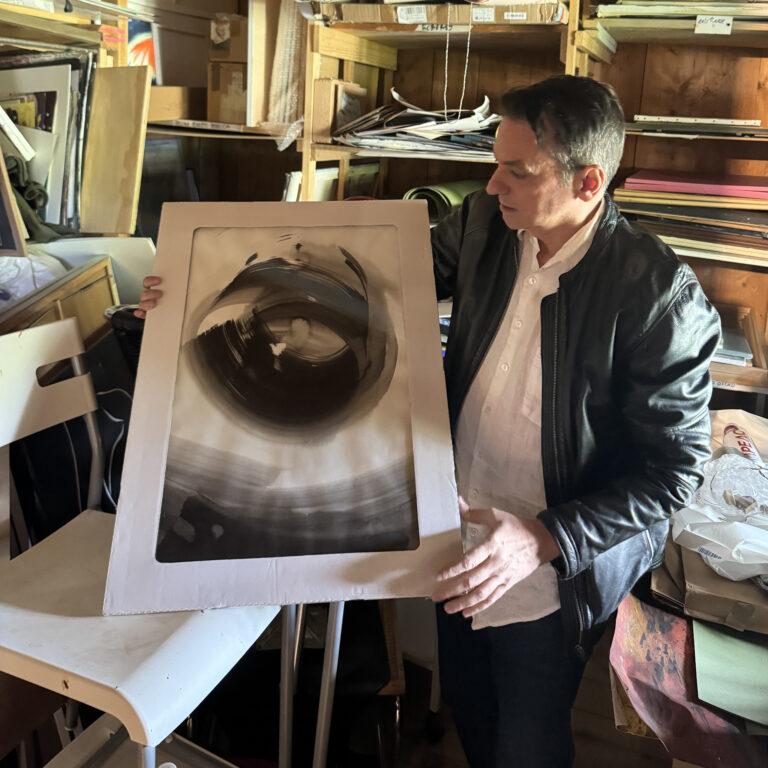 Ein Mann in Lederjacke hält ein gerahmtes, abstraktes Schwarz-Weiss-Kunstwerk in einem Atelier.
