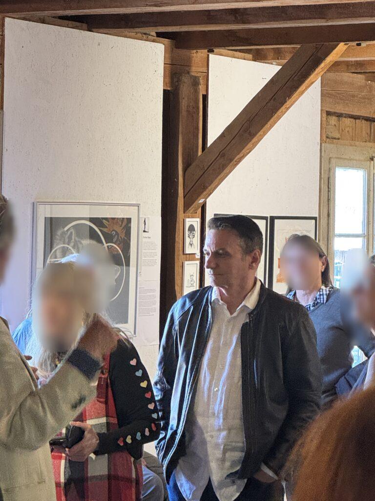 Ein Mann in dunkler Jacke und weißem Hemd steht in einer Galerie vor gerahmten Kunstwerken.