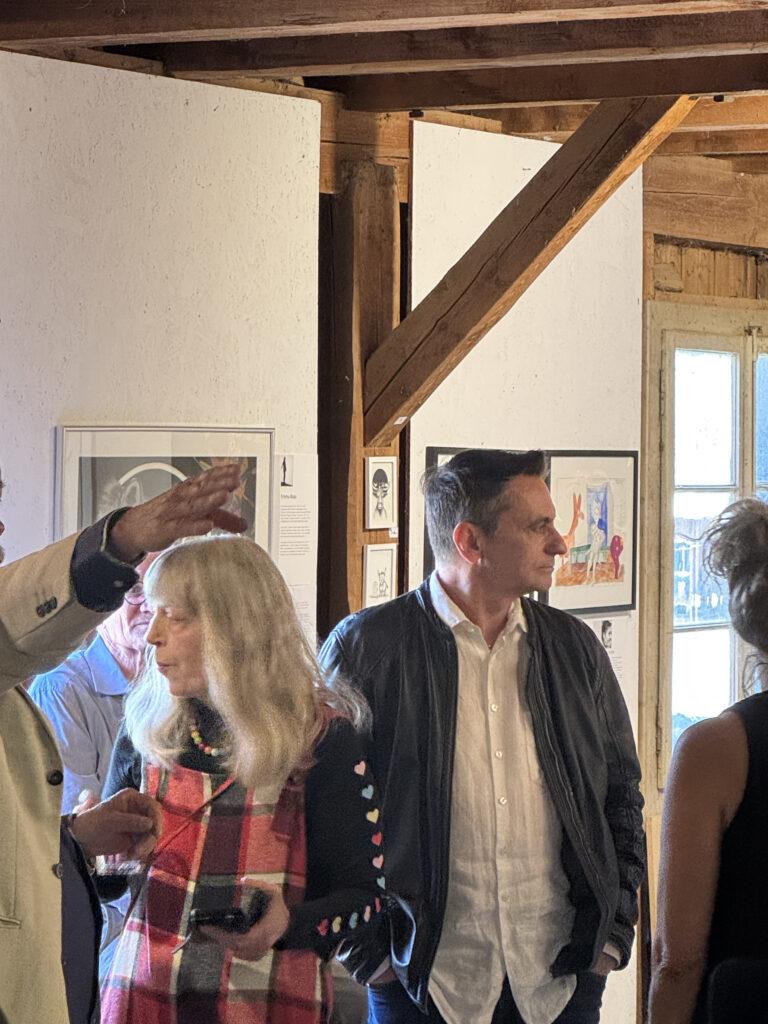Personen stehen in einem hellen Raum mit Holzbalken vor gerahmten Kunstwerken an einer weißen Wand.